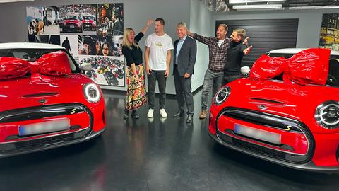Eine Gruppe Personen, darunter Tobi, Tanja und Gewinner Justin, stehen in einem Autohaus zwischen zwei roten MINI Cooper.