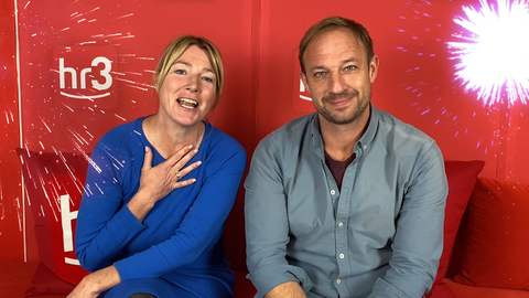 Tobi und Tanja sitzen lachend vor der roten hr3 Studiowand