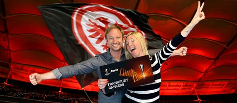 Tobi und Tanja halten vor dem Hintergrund der Eintracht-Fahne aufgeregt einen Ticketgutschein in die Kamera