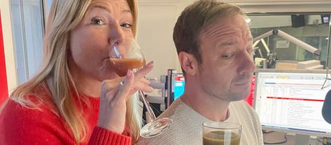 Tanja hält lächelnd ein Saftglas in die Kamera. Tobi schaut sein Glas mit grünem Saft skeptisch an.